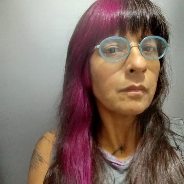 A portrait image of a woman with long dark hair and a magenta streak through her blunt bangs. She wears blue-framed glasses and stares at the camera with a serious expression. Both the background and her sleeveless T-shirt are grey. On her shoulder and neck we can see the hint of two graphic black tattoos. 