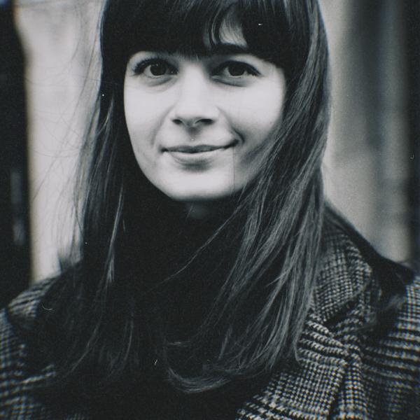 A black and white portrait image of a woman with long dark hair and blunt bangs. The image is a high-contrast photograph, with a light blurring effect on the subject's outline. The subject smiles to one side and has dark eyes; they wear a houndstooth blazer over a dark top. 