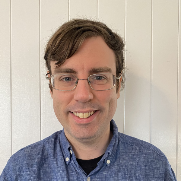 A photographic portrait of a man shot against a wall of painted white wood panelling. He smiles at the camera and wears wire-rimmed glasses and a blue button-down shirt. He has light brown hair parted to one side. 