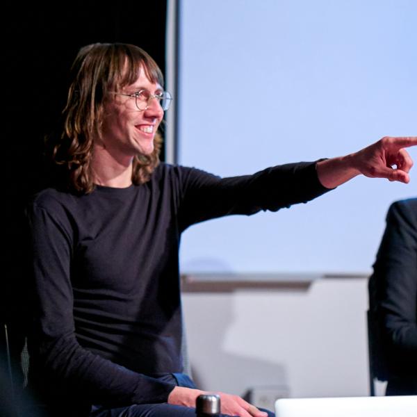 A photograph shows a person in partial profile sitting at a table. They are pointing off camera and sit beside anotehr person, whose face is cropped out by the camera. The subject has long, light brown hair with short bangs. They wear wire-rimmed glasses and a long-sleeved black T-shirt. There is a strong light source and the cropped appearance of camera with a glowing green light. 