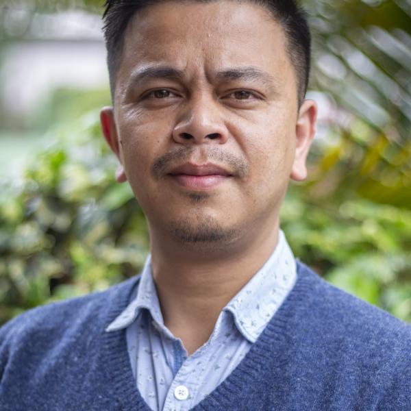 A portrait photograph of a man squinting slightly into the camera. He is wearing a blue collared shirt underneath a blue V-neck sweater. He has short black hair and dark eyes. The background shows a blue sky and tropical plants, out of focus. 