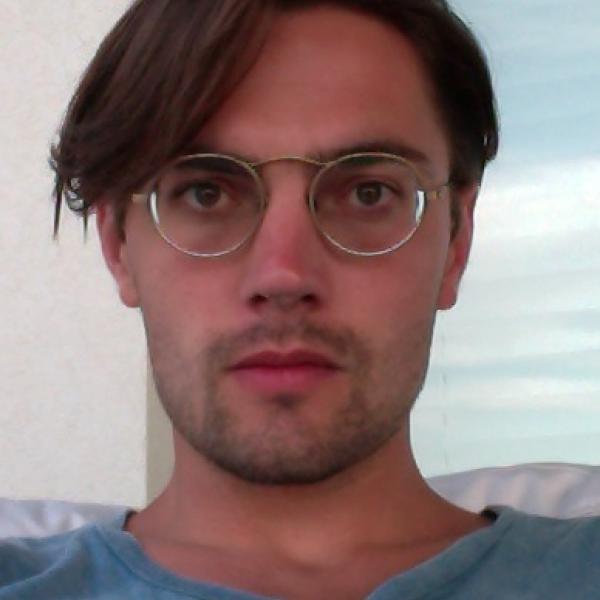 A portrait photo of a man staring into the camera, shot from the shoulders collarbone up. They have short brown hair swept to one side, and wear a blue T-shirt with wire-rimmed glasses. They have brown eyes and stubble on their face.  