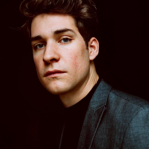 A portrait photograph of a young man standing in front of black background. They are wearing a dark sports jacket or blazer, a black shirt, and have short dark brown hair that is lightly styled.