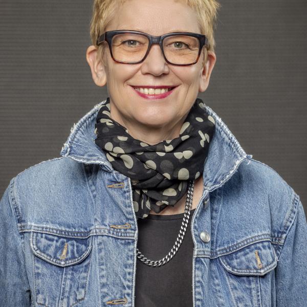 A colour photograph of a woman with short blonde hair, dark glasses, a jean jacket with the collar popped up, a dark patterned scarg, a chain necklace, and a dark grey shirt. She is standing in front of a dark grey wall and smiling. 