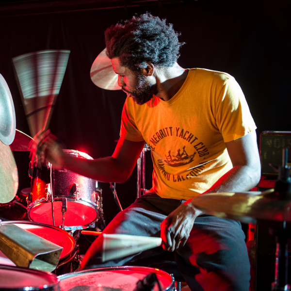 A colourful photograph of a man sitting at a drum kit. He wears a yellow t-shirt and is shot in partial profile, the drums in front of him. He is pictured in action, a drumstick blurred in motion. He is lit with strong red and white light sources. 