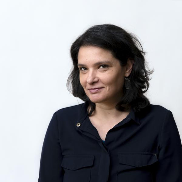 A woman in a black button-down shirt is photographed from the elbows up against a white background. She has shoulder length black hair and dark eyes wears dark sculptural earrings. She smiles and looks into the camera. 