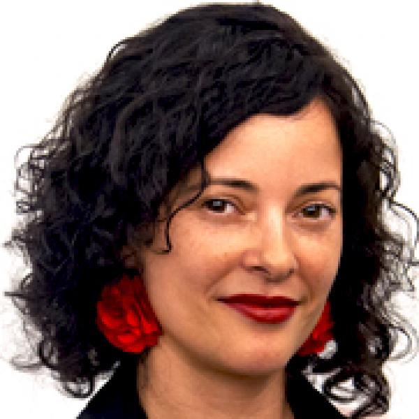 A portait headshot photo of a women with shoulder-length curly dark hair, large hanging red earrings, and red lipstick. She is wearing a black collared shirt or jacket that is mostly cropped out of the photo and standing in front of a white background.