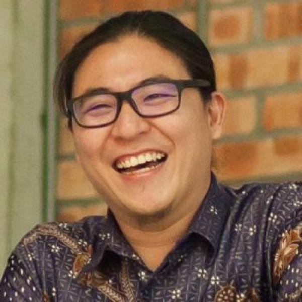 A photo of a person with black hear and horn-rimmed glasses who is laughing in a room with exposed brick walls. They are wearing a dark purple-grey patterned collared shirt.