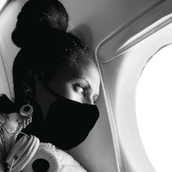 A black and white photograph of a woman with dark hair who is sitting in an airplane on a window seat looking out. She is wearing a black mask over her nose and mouth, dangling earrings, and large headphones.