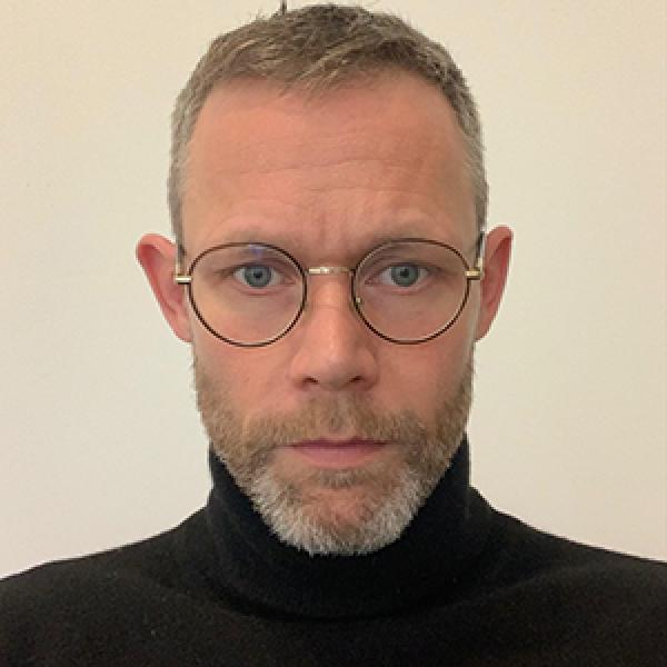 A headshot photo of a man with round wireframe glasses, a short beard, short blond hair, and a black turtle neck. He is standing in front of a plain beige background.