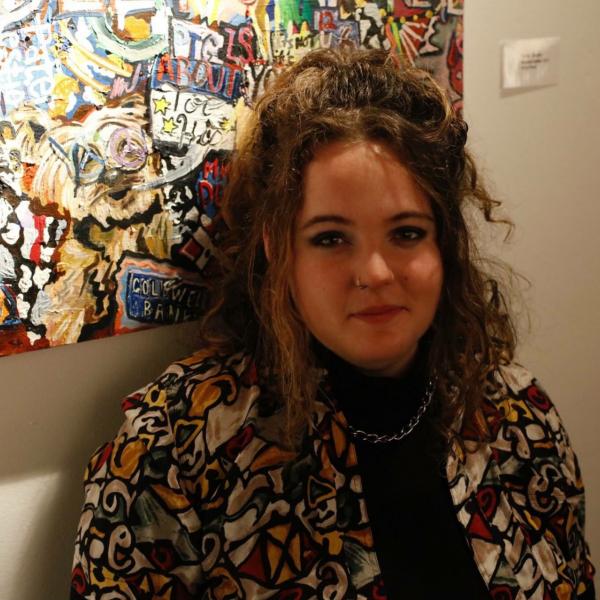 A photograph of a woman standing in front of an abstract painting. She looks into the camera and is standing in partial shadow. She has curly hair tied up on her head, a nose piercing, and wears a colourful patterned shirt over a black turtleneck with a thick chain necklace. 