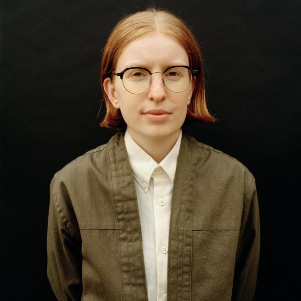 A striking photograph of a person standing against a black background. They look intently into the camera and wear wire-framed glasses with a white button-down shirt and an army green jacket. They have shoulder length red hair, parted in the middle. 