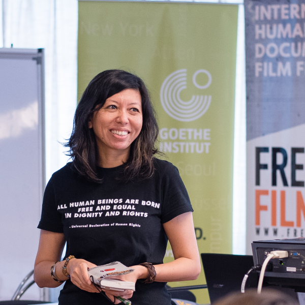 A photograph of a woman standing in front of signs and a projector. She smiles off camera and holds a book in her hands. She has shoulder length black hair and wears a black T-shirt that reads "ALL HUMAN BEINGS ARE BORN FREE AND EQUAL IN DIGNITY AND RIGHTS." The signs in the background read: "Human Rights Documentary Film Festival" and "Freedom Film Fest."