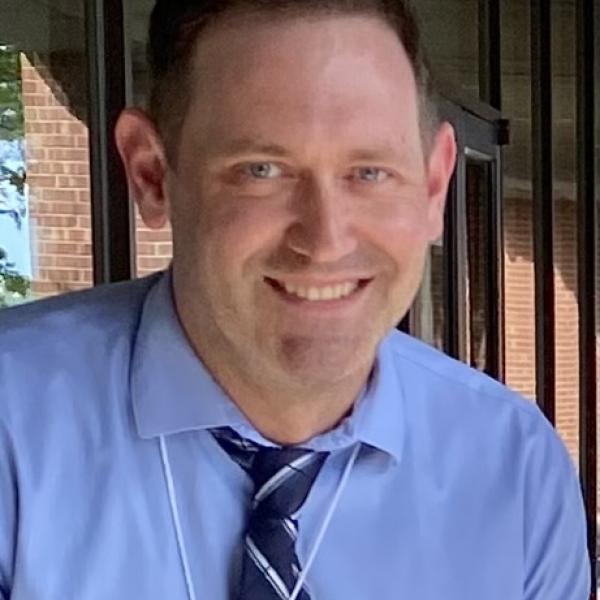 A man in a blue shirt and a striped navy and white tie smiles at the camera. He has blue eyes and close-cropped light brown hair. In the background are windows reflecting a brick walls and a bright blue sky.