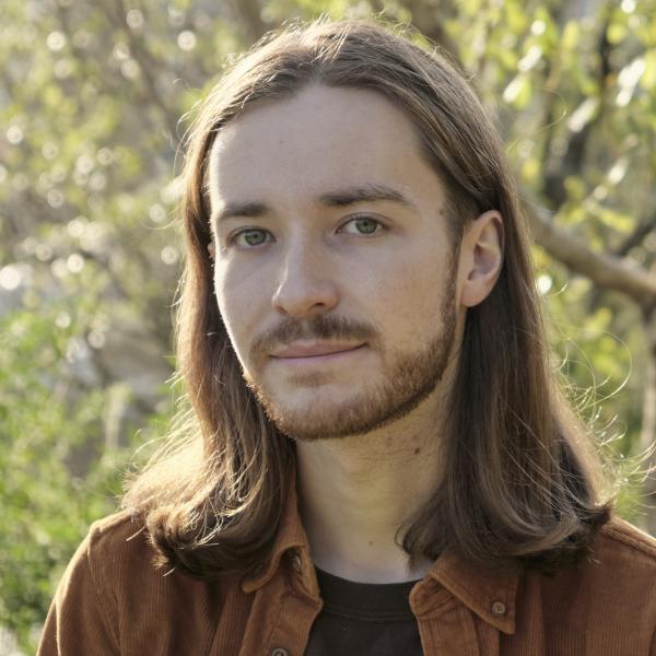 A colour portrait photograph of a young man with shoulder-length light brown straight hair and a light beard. They are slightly smiling and wearing a brown collared shirt and a dark t-shirt. they are standing outside in a forest on a sunny day.