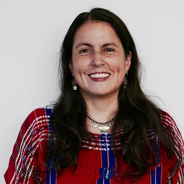 A portrait photograph of a woman standing against a white background. She smiles into the camera and wears a colourful, red and blue top. She wears an ornate silver necklace and silver earrings and has long dark hair. She is shot from the chest up. 