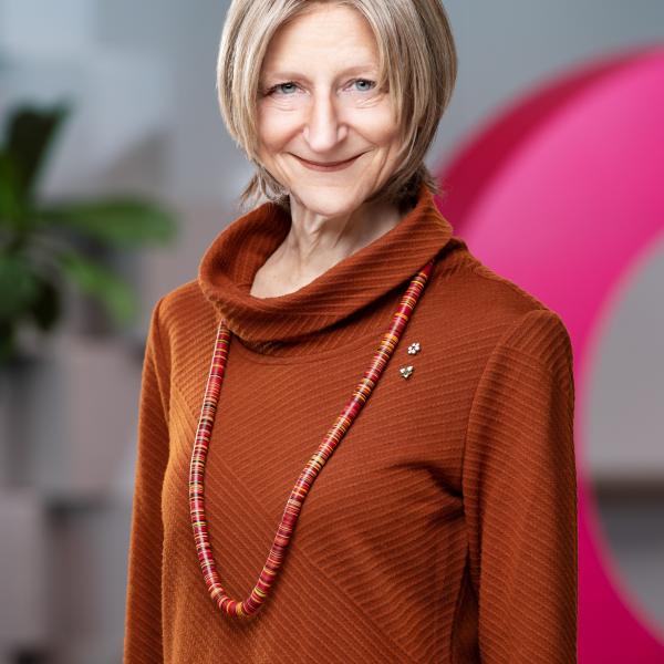 A professional looking portrait photograph of a woman standing against a graphic, grey background with a large circular pink shape. She has short, blonde hair chopped at the chin and wears a burnt orange sweater with a colourful beaded necklace. She smiles at the camera with her mouth closed. 