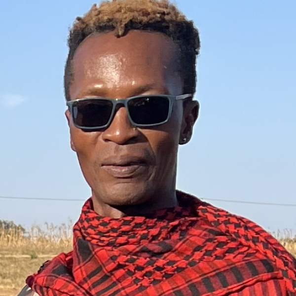 A portrait image of a man shot against a light blue sky. He has short black hair with blonde accents, and wears dark grey sunglasses. He wears a red patterned scarf and a stud earring in his left ear. It appears to be a bright sunny day. 