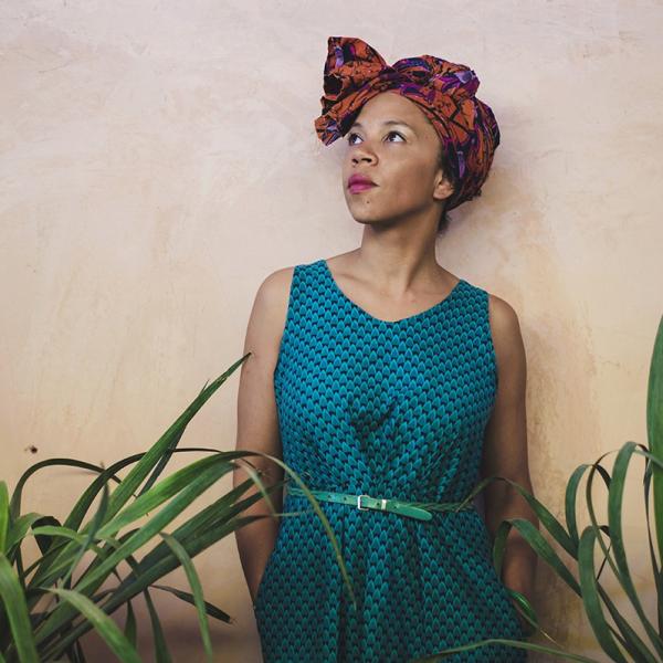 A photograph of a woman standing against a textured, clay wall. She looks up and to the side and is shot in partial profile. She wears a colourful headscarf of oranges and purples, and a blue patterned dress with a thin teal belt. She has on a bright pink lipstick. Leafy plants sit in the foreground. 