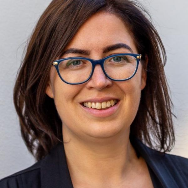 A portrait photograph of a woman smiling into the camera. She wears a white T-shirt underneath a navy blue blazer. She has shoulder-length red-brown hair and blue-framed glasses. She is shot against a light grey background. 