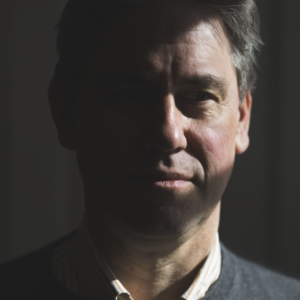 A close-up photographic portrait of a man's face. He is shot almost entirely in shadow, with sharp rays of light illuminating parts of his face. He looks off to the side and has short dark grey hair. He wears a collared shirt beneath a V-neck sweater.  