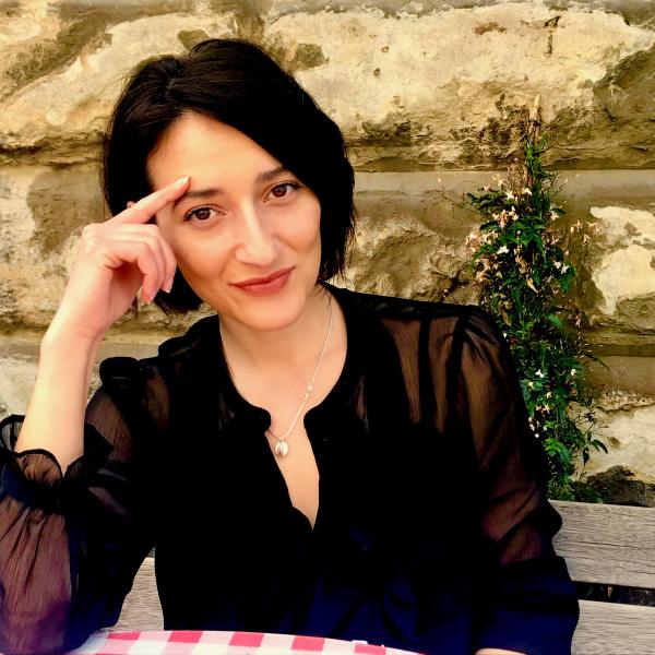 A portrait photograph of a woman sitting at a table in front of a stone wall. She has straight, chin-length black hair and rests her face on her hand. She wears a sheer black blouse with a silver necklace. She looks intently at the camera with a slight smile on her face. 