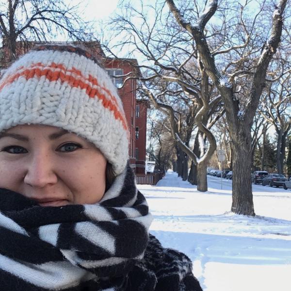 A photograph of a woman shot against a snowy background. She wears a knitted hat and a black and white striped scarf that partially obstructs her chin. She smiles at the camera. In the background are snow covered trees and red brick buildings. 