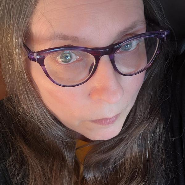 A close-up of a woman's face, shot from above. She looks into the camera with a serious expression on her face, and wears purple-framed glasses. The subject wears a black T-shirt and has blue eyes and light brown hair parted in the middle. 