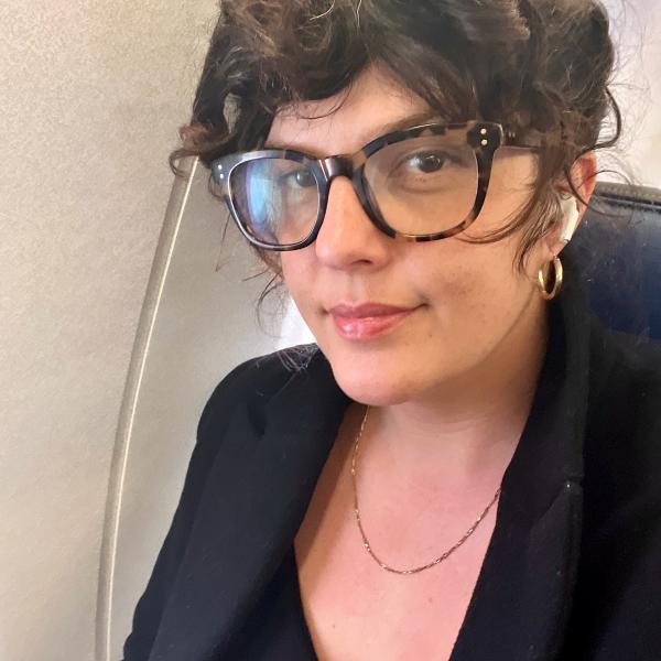 A portrait image of a woman with dark curly hair wearing tortoiseshell glasses. She has AirPods in and wears a black blazer over a black T-shirt, with a gold chain around her neck and small gold hoops in her ears. She smiles slightly at the camera. She appears to be in an airplane seat. 