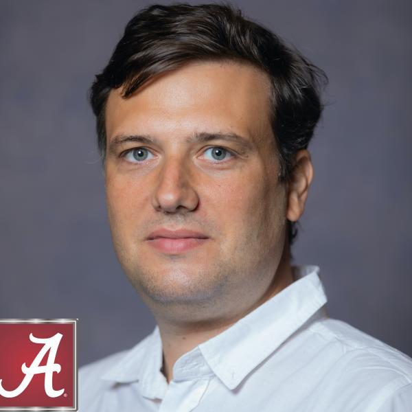 A close-up photographic portrait of a man looking into the camera, wearing a white collared shirt. He is shot against a dark grey background and has blue eyes and short, light brown hair. 