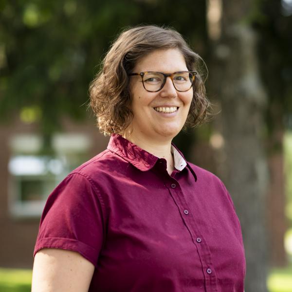 A portrait of a person shot from the waist up, in front of a blurred background of trees and brick buildings. They wear a short-sleeved magenta button down shirt with tortoiseshell glasses and have curly brown hair to their chin, parted to the side. They smile brightly at the camera. 