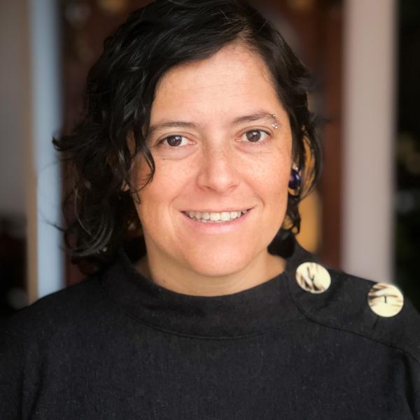 A portrait photograph of a woman smiling at the camera. The image is well-lit and the subject is shot from the shoulders up. She wears a sweater with large buttons on the shoulders and has short, dark hair with a piercing on one eyebrow. 