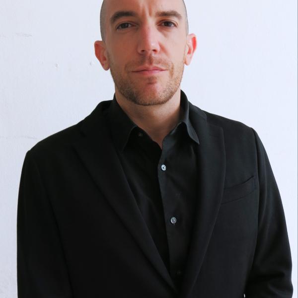 A photograph of a man shot from the waist up in portrait style. He wears a black blazer over a black shirt and looks into the camera. He has a buzzed haircut and a short beard. He is shot against a bright white background. 