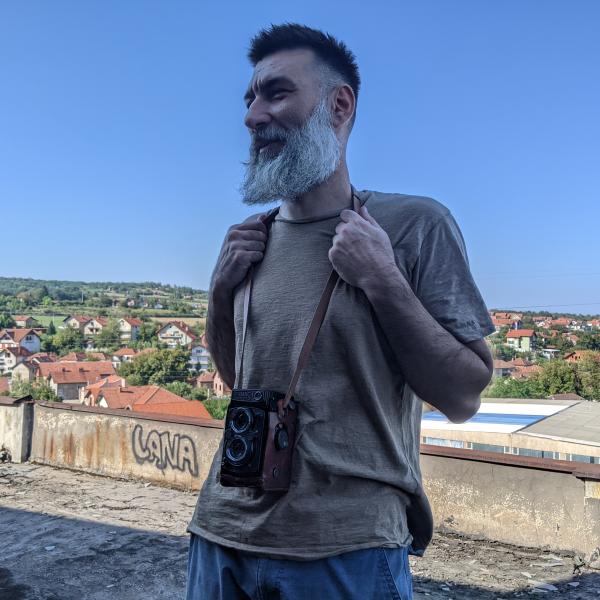 A photograph of a man standing on a road with a camera around his neck. He is shot in 3/4 profile and has a smile on his face. He wears a casual T-shirt and jeans, has a long white beard and short black hair. The background shows rolling hills covered in houses and buildings with terracotta roofs. 