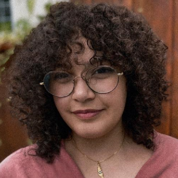 A close-up photographic portrait of a woman with curly hair and wire-rim glasses. She wears a dusty rose v-neck top with a gold necklace. The blurred background shows a wooden fence and plants creeping up a white wall. 