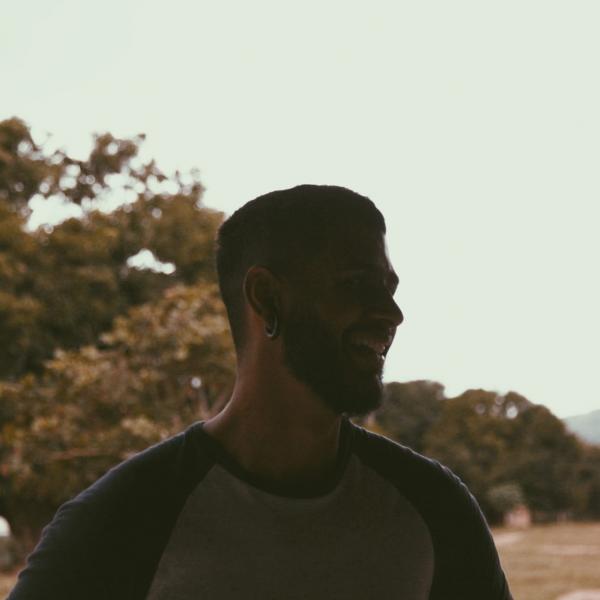 A photgraph of a man shot in partial profile in front of a treed background. He is lit from behind and smiles. He wears a baseball T-shirt and has short, dark hair and a thick beard. 