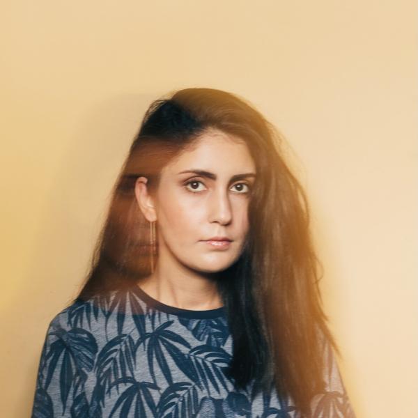 An artistic portrait photograph of a woman with long dark hair parted to one side. The background is a beige colour, and there is an interesting blurring effect over her image. She has dark eyes and wears a blue T-shirt with a pattern of palm fronds on it. She has one long earring. 