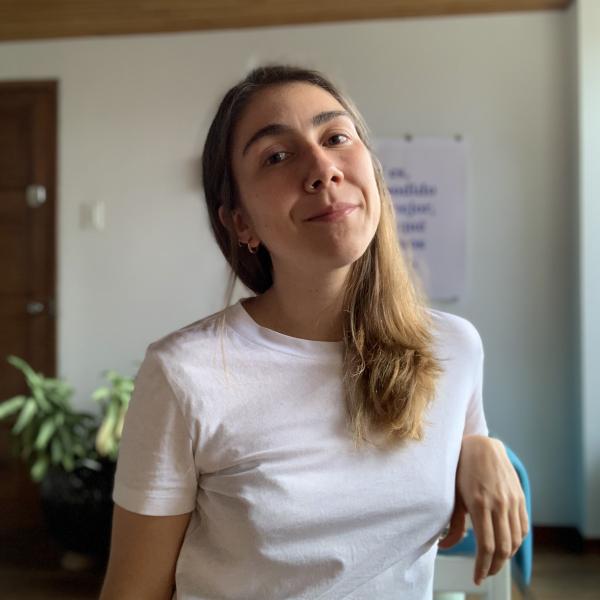 A photograph of a woman in a white T-shirt sitting in a chair, shot head on. She has long, dark blonde hair and looks into the camera. The blurred background shows a wooden ceiling and white walls with a large plant in the corner of the frame. 