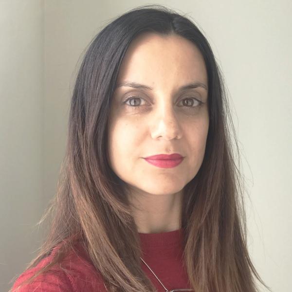 A portrait photograph of a woman looking into the camera, wearing a red shirt. She has dark eyes and long dark hair with blonde colouring towards the ends. She is shot against a light grey background and wears red lipstick that matches her shirt. Around her neck is a sculptural silver necklace. 