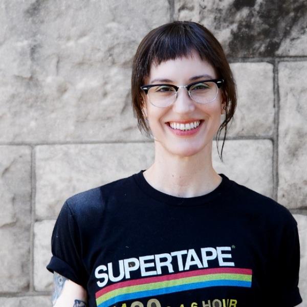 A portrait photograph of a woman against a grey stone brick background. She has short dark hair with short bangs and wears horn-rimmed glasses. She smiles brightly at the camera and wears a black T-shirt with a rainbow graphic on it and the word "SUPERTAPE."