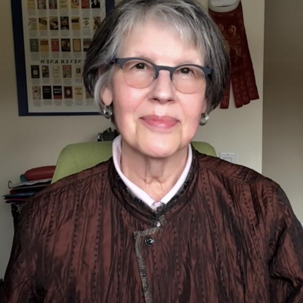 A portrait image of a woman seated in a green office chair. The subject has short, straight grey and black hair and wears rounded glasses with small silver hoop earrings. She smiles with her mouth closed and wears a textured burgundy jacket. In the background a graphic poster hangs on the wall, along with a red textile. 