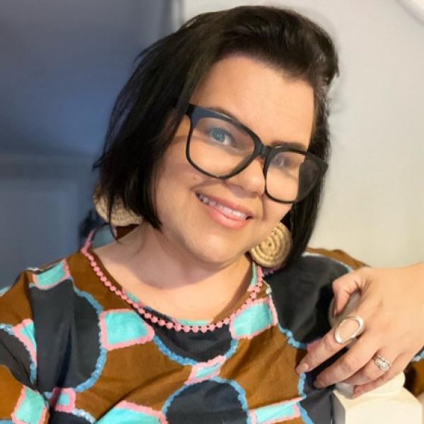 A photographic portrait of a woman with chin-length dark hair and thick black-framed glasses, wearing a bright patterned shirt and straw earrings. She shows her hand, which has silver rings on it, and smiles with her head tilted to one side. 