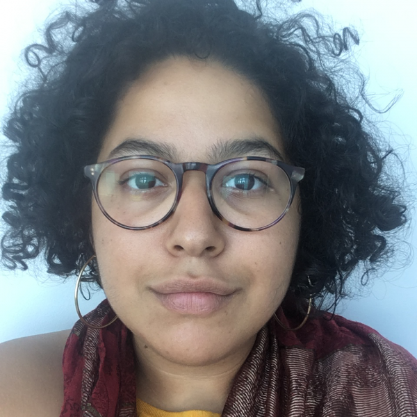 A portrait image of a woman looking directly into the camera. They have dark, curly hair and wear horn-rimmed glasses. They wear a yellow shirt and a burgundy, patterned scarf as well as hoop earrings. The background is a bright white. 