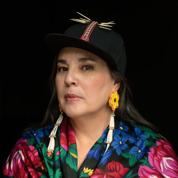 A colour photograph of a woman in a floral shirt with a black baseball cap and long colourful dangling earrings