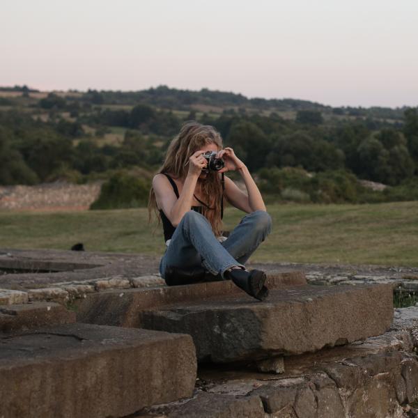 A photograph of a woman sitting on a wall of large stones, with an analog camera to her face. Rolling hills in the background are covered in short, dark green trees. The woman has long, dark blonde hair and wears blue jeans, black boots, and a black tank top. 