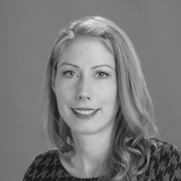 A greyscale photo of a woman with blond shoulder-length hair standing in front of a plain background.