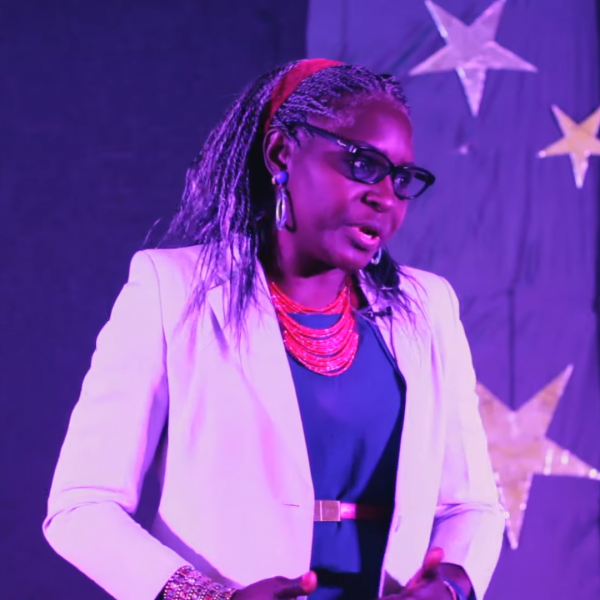 A screenshot of a woman in a white blazer, wiith a pink necklace, a blue blouse, and greying, braided hair that is tied back with a red ribbon. She is standing on a stage with a purple piece of fabric with shiny cut-out stars attached to it.