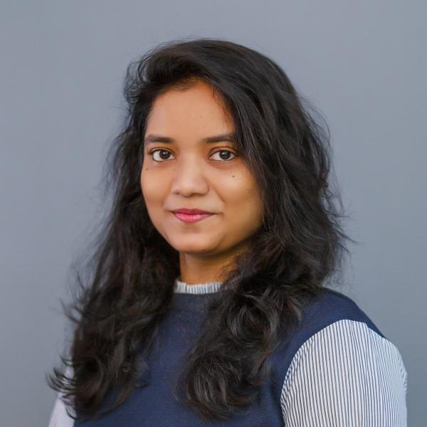 A portrait photo of a young woman with long dark hair who is standing in front of a plain grey background. She is wearing a navy blue vest and a blue and white striped shirt underneath.