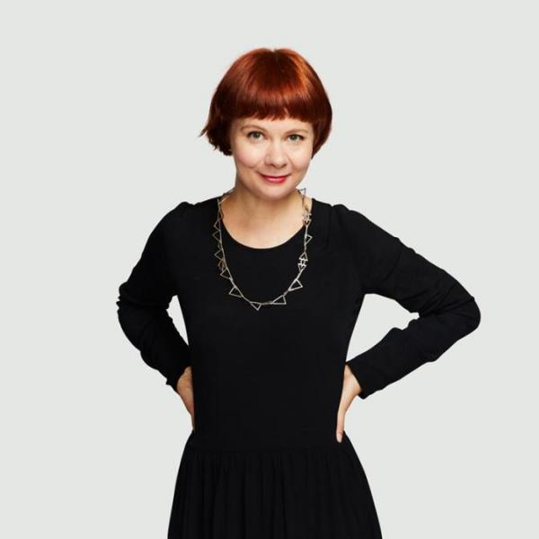 A portrait photo of a woman standing against  aplan white background. She is wearing a black dress and a long necklace and has short red hair styled in a bob. She has her hands on her hips and is slightly smiling.