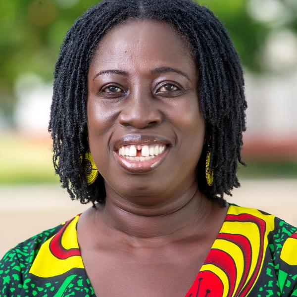 A portrait photo of a smiling woman standing outside on a sunny day. She has shoulder-length, dark, braided hair; large yellow-gold hoop earrings; and a colourful patterned shirt.